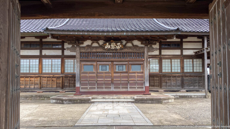 Wooden Buddhist temple hall with tiled roof, central doors, lattice windows, carved plaque, and stone path.