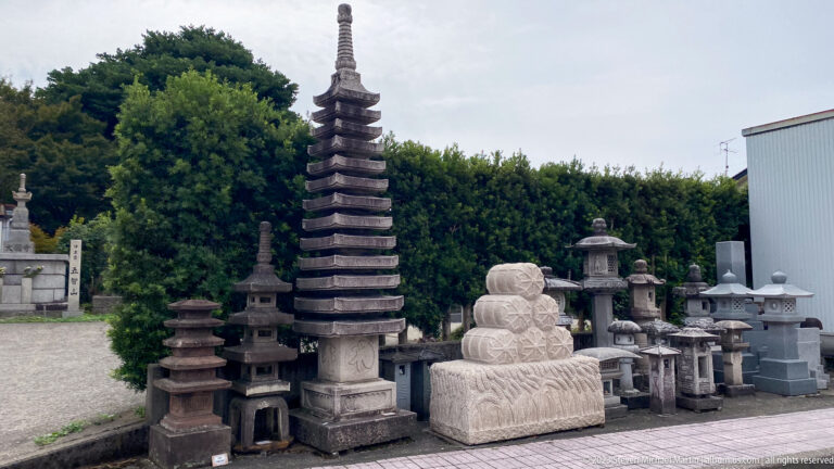 Multi-tiered stone pagoda surrounded by stone lanterns, carved bases, and memorial objects in an outdoor display yard.