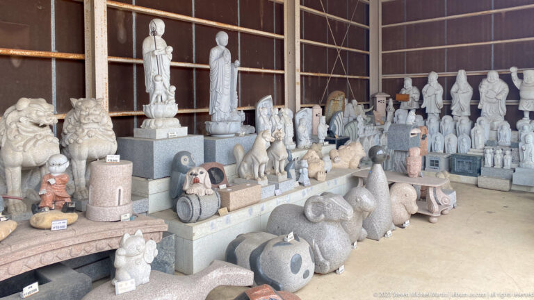 Open workshop display filled with stone Buddhist figures, guardian lions, animals, lanterns, and memorial carvings.
