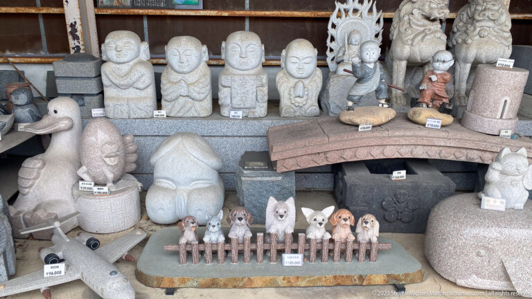 Stone display with smiling Jizō-like figures, guardian animals, pet memorial sculptures, and small decorative carvings.