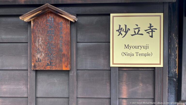 Bilingual Myōryū-ji “Ninja Temple” sign mounted beside an older vertical wooden plaque on a dark timber wall.
