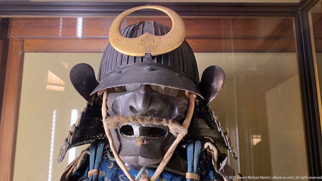 Close view of samurai helmet with broad gold crest, dark face mask, cords, blue lacing, and shoulder armor.