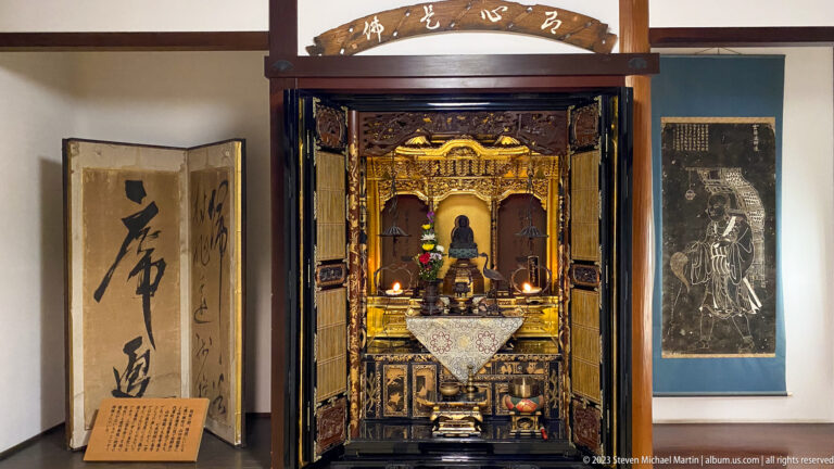 Open gilded butsudan with seated Buddha, flowers, candles, calligraphy screen at left, and devotional image at right.