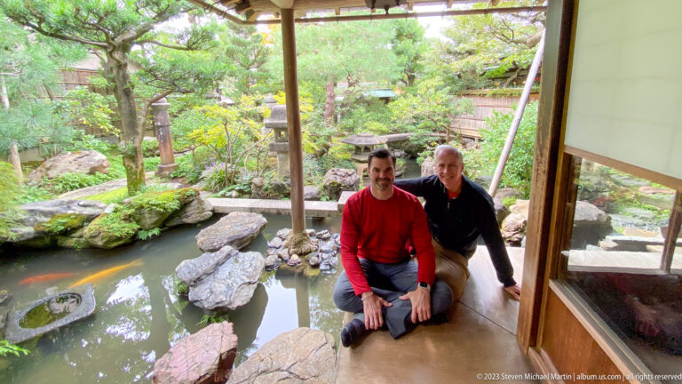Two male visitors seated on a wooden veranda beside a pond with stepping stones, moss, shrubs, and lanterns.