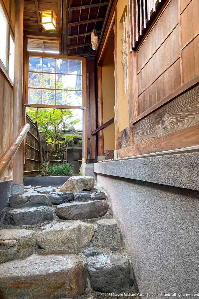 Narrow stone-paved passage with handrail beside timber siding, plaster foundation, and a small garden beyond.