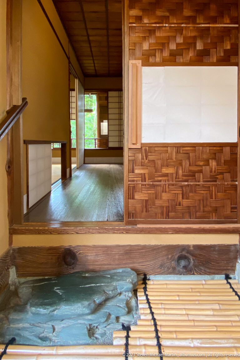Raised wooden room with woven wall panels, white plaster, sliding screens, bamboo slats, and dark floor opening.