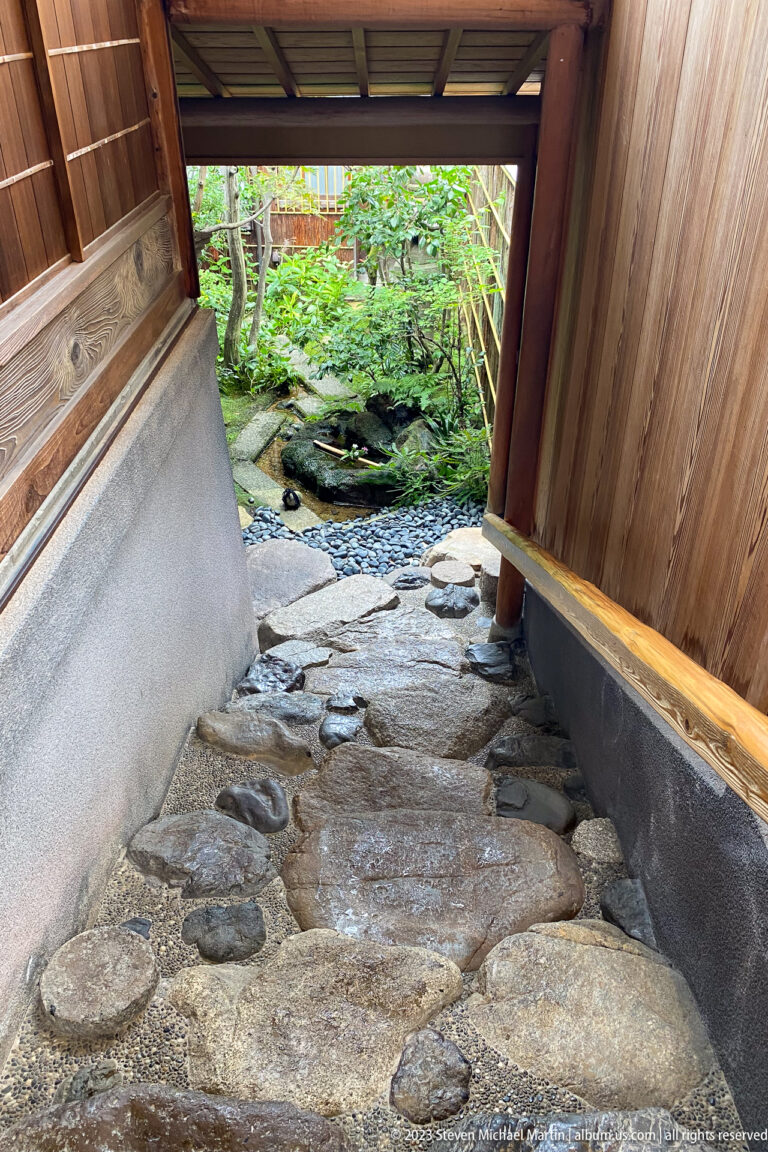 Covered stone passage between wooden walls leading toward a green garden and water beyond.