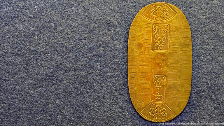 Single elongated gold-toned Japanese currency piece with floral emblems and vertical inscriptions.