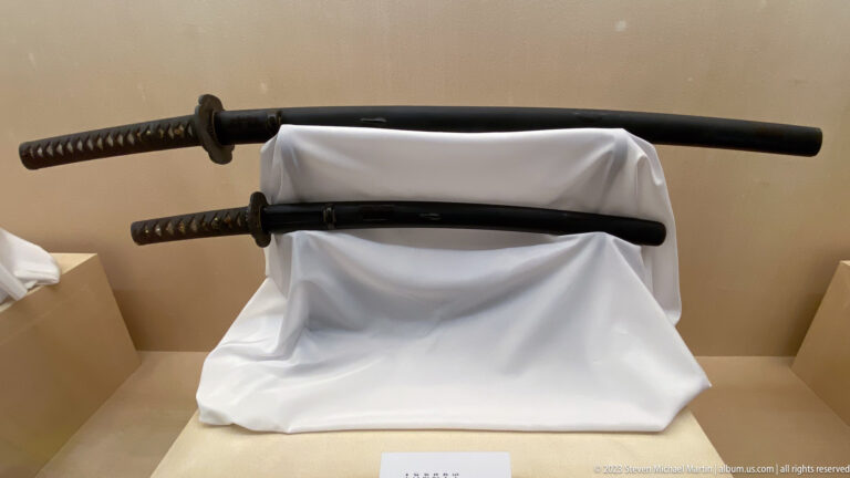 Two sheathed Japanese swords on a white-draped stand, one longer above and one shorter below.