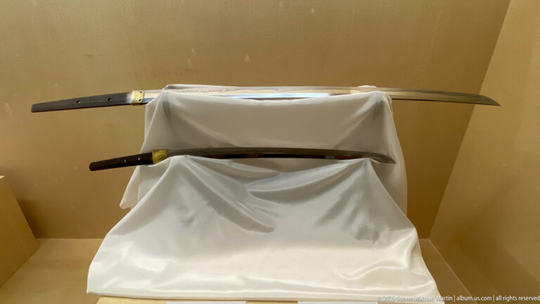Two unsheathed curved Japanese swords displayed above dark scabbards on a white-draped stand.