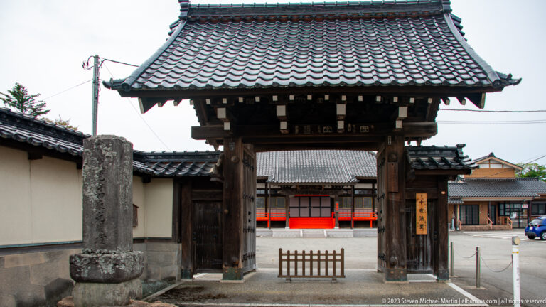 Frontal view of a timber Buddhist temple gate with tiled roof, plaster walls, and red courtyard elements beyond.