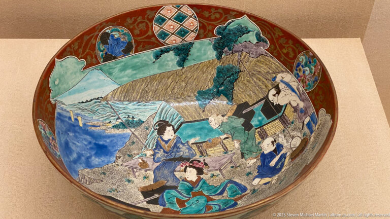 Large polychrome ceramic bowl painted with figures, water, thatched buildings, mountains, and patterned rim medallions.