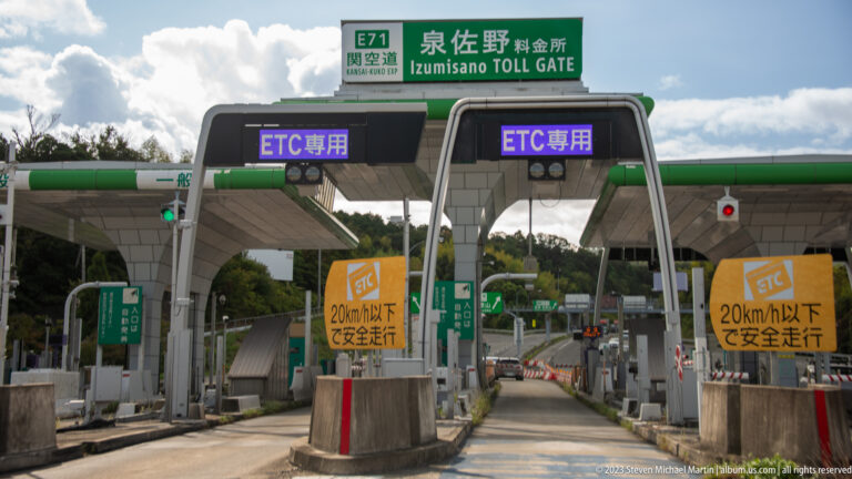 Izumisano toll plaza with green overhead signs, ETC lanes, booths, and vehicles beneath a blue sky.