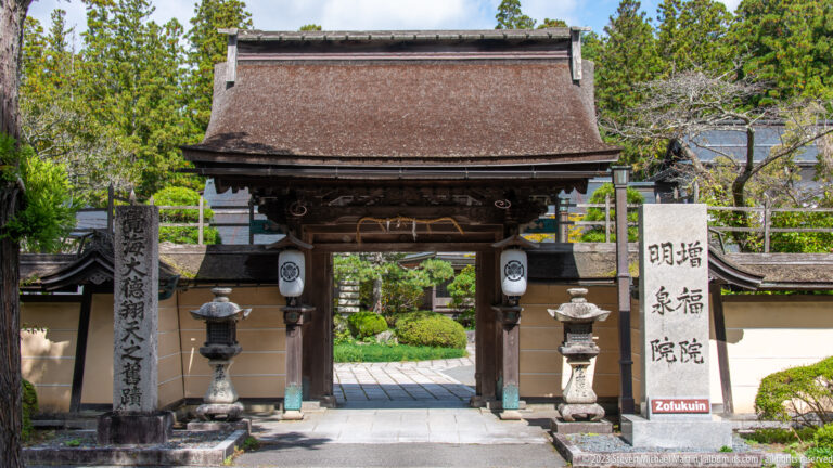 Wooden Zofukuin gate with hanging lanterns, stone lanterns, white walls, and a garden beyond.