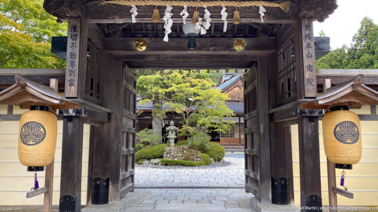 Wooden Kongōbu-ji gate with crest lanterns framing a landscaped inner courtyard.