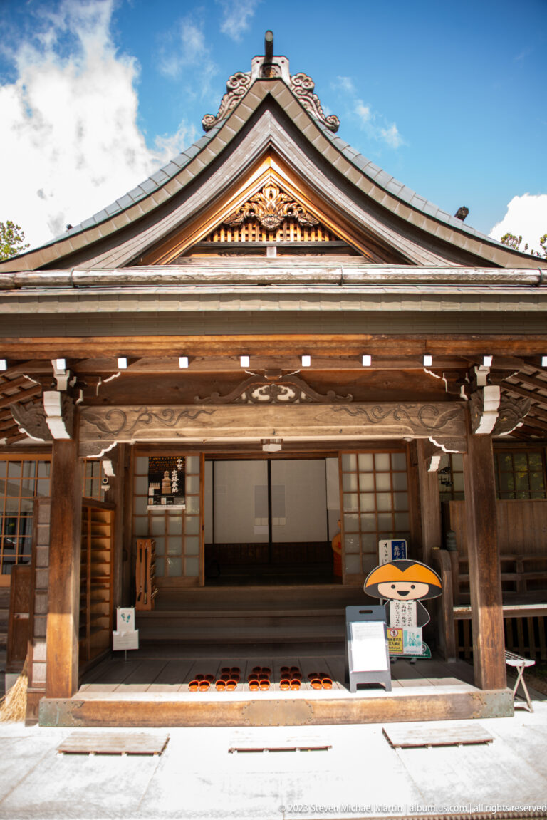 Traditional wooden building with open veranda, sliding doors, carved gable, floor mats, and a mascot figure.