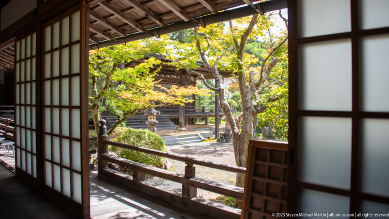 Wooden temple corridor with shoji screens opening toward a sunlit garden, shrubs, and stone lanterns.