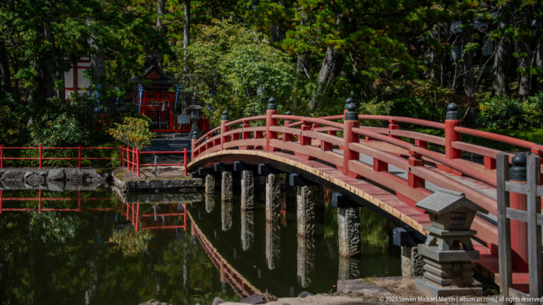 Vermilion arched bridge reflected in a pond, with trees and a small red shrine beyond.