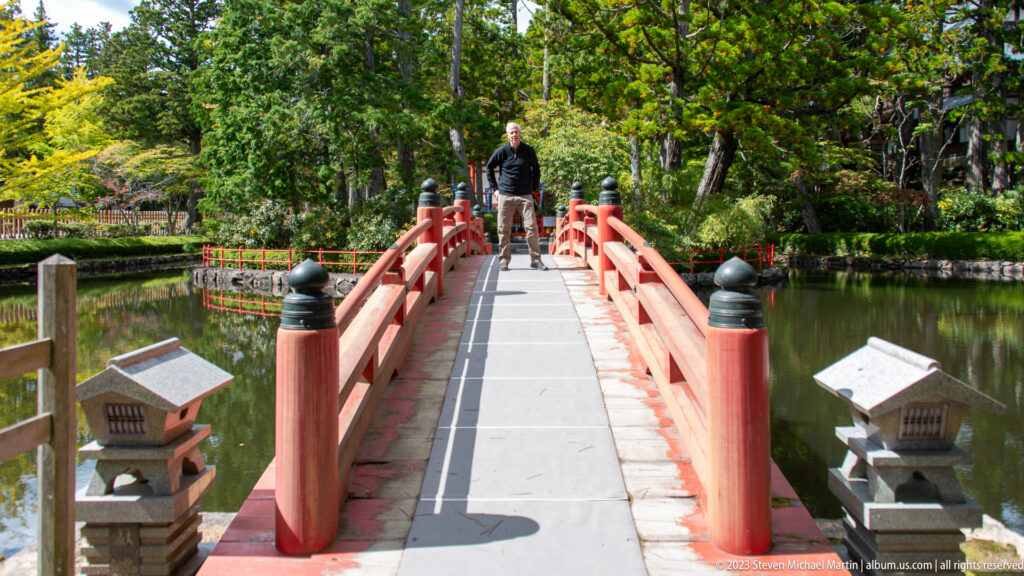 Visitor standing at the center of a red arched bridge over a pond, flanked by stone lanterns.