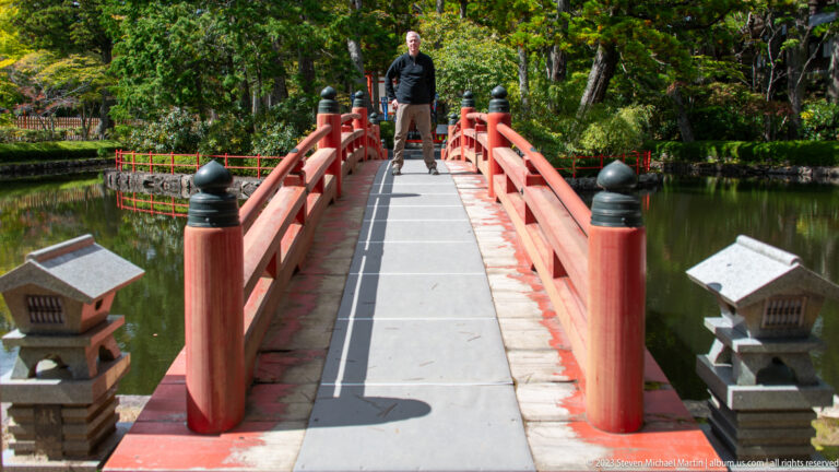 Visitor standing on a red arched bridge viewed from the entrance between two stone lanterns.