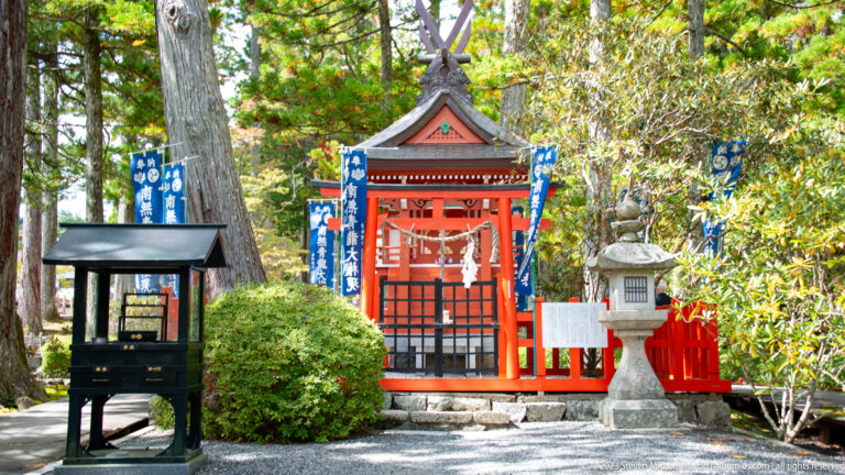Small red shrine-like structure among trees with blue banners, railings, and a dark offering fixture.