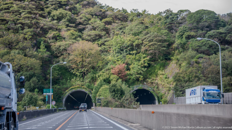Two highway tunnels open into a densely forested hillside as cars and a truck travel along the road.