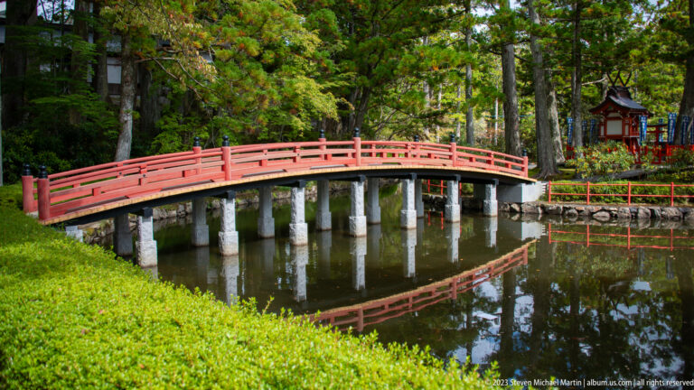 Side view of a red arched bridge on stone piers reflected in a still pond.