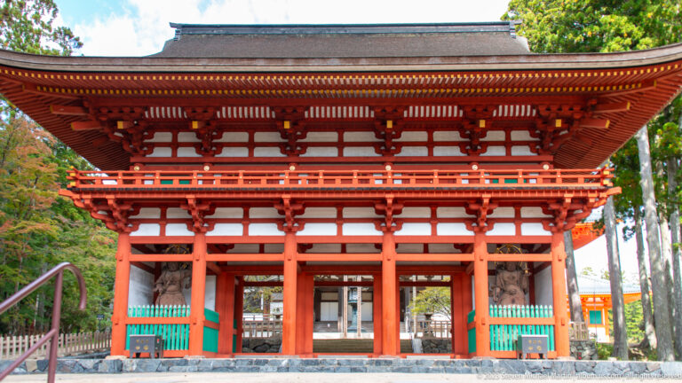 Large vermilion two-story Buddhist gate with white panels, tiled roof, and guardian statues in side bays.