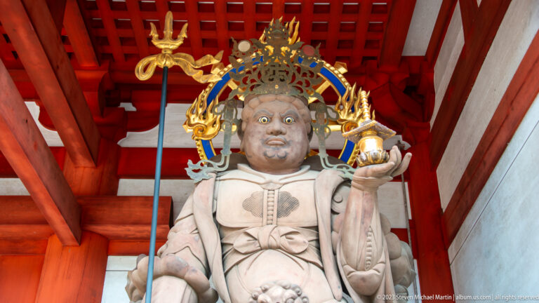 Painted Buddhist guardian statue with bulging eyes, ornate crown, long staff, and raised hand.