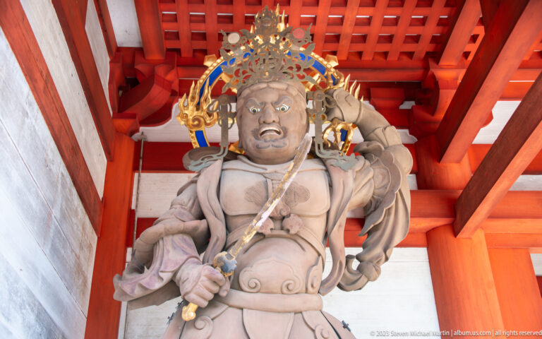 Muscular Buddhist guardian statue with raised arm, fierce face, and weapon beneath red gate beams.