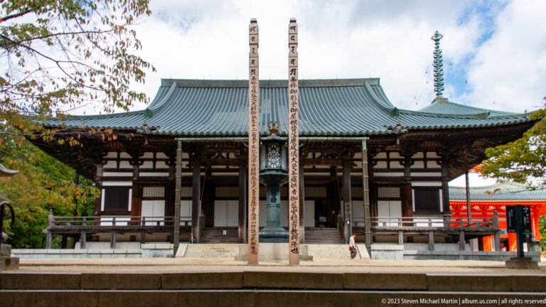 Large dark-timber Buddhist hall with broad tiled roof and two tall inscribed pillars in front.