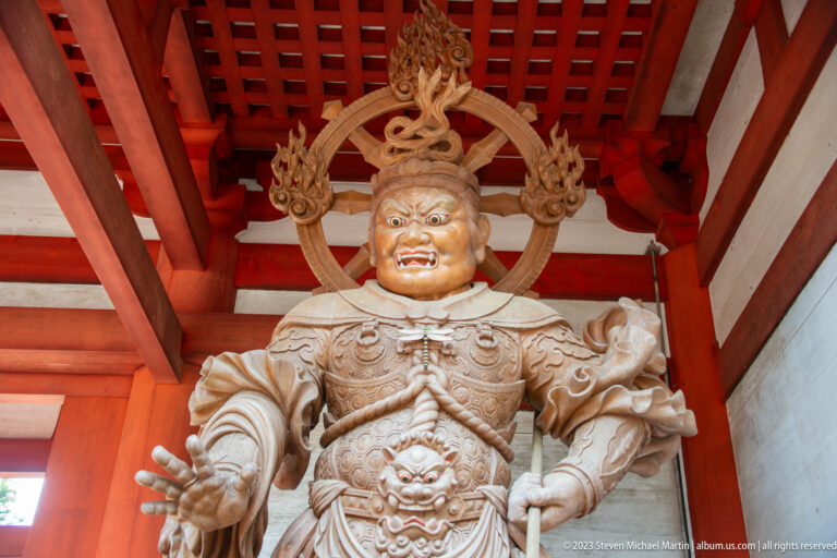 Armored Buddhist guardian with fierce face, extended hand, ornate crown, halo, and flowing sash.