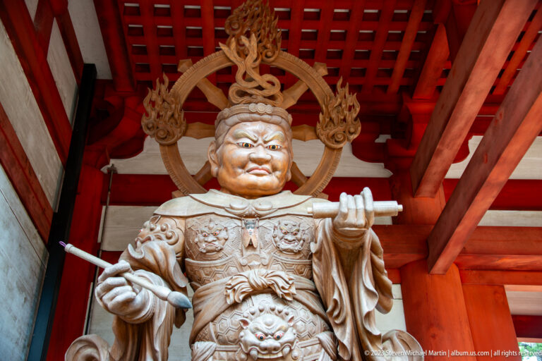 Front-facing armored Buddhist guardian with raised hand, straight attribute, fierce face, and flame-like halo.