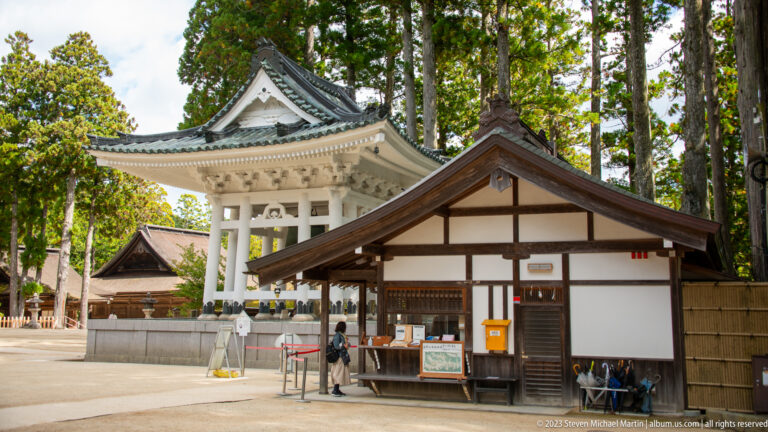 White open-sided tiled structure beside a small wooden visitor office among tall trees.