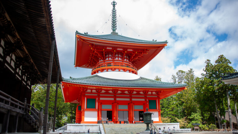 Vermilion Konpon Daitō pagoda with broad eaves and tall finial beneath a partly cloudy sky.