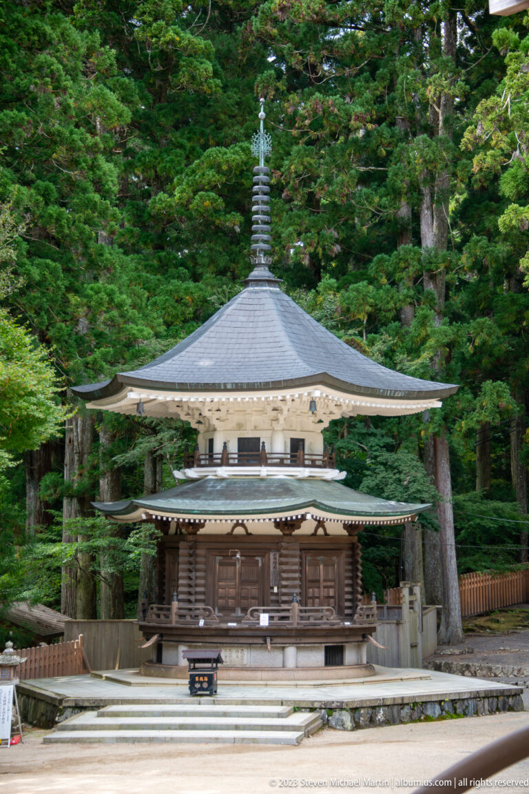 Small dark-wood Buddhist hall with a low pyramidal roof and finial among tall cedar trees.