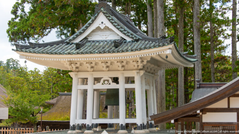 White open-sided bell pavilion with a suspended bronze bell and gray tiled roof.