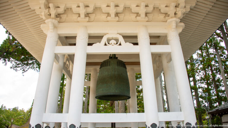 Large bronze temple bell hanging inside a white open-sided timber pavilion, viewed from below.