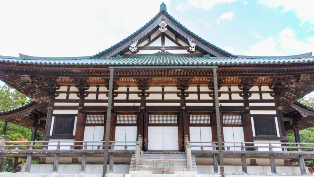 Symmetrical dark-timber Buddhist hall with white panels, tiled gable roof, veranda, and central steps.