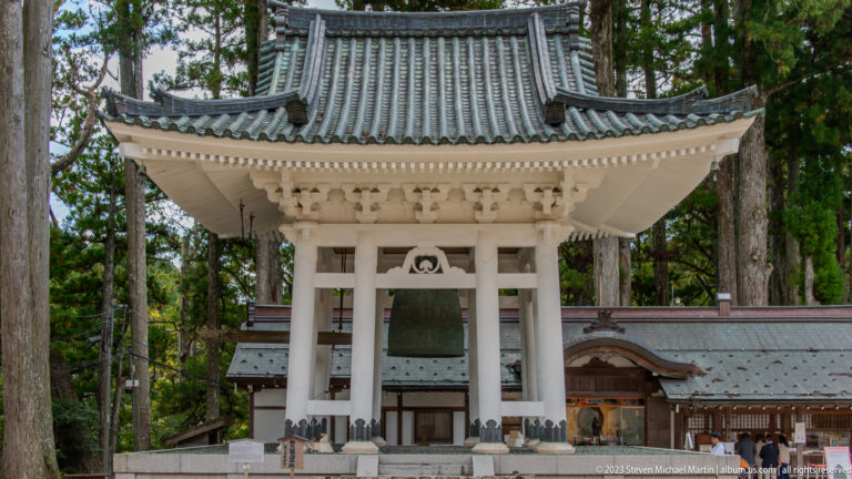 White bell pavilion with visible bronze bell, tiled roof, nearby temple buildings, and tall cedars.