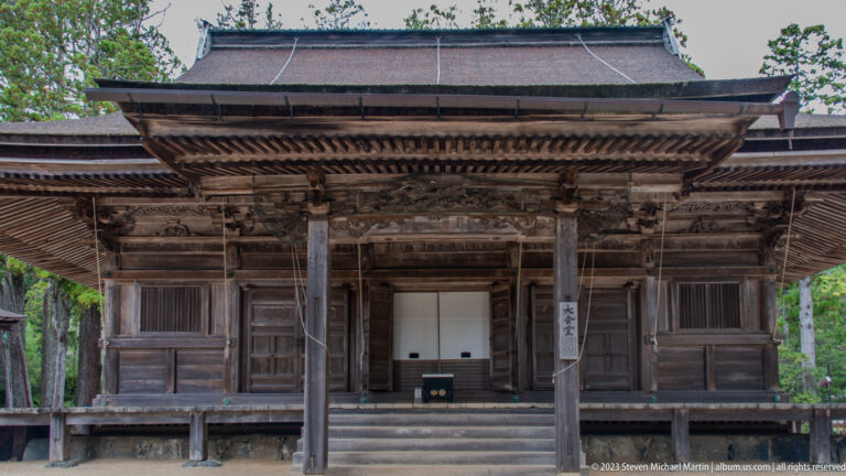 Weathered dark-wood Buddhist hall with deep veranda, carved brackets, and shuttered front bays.