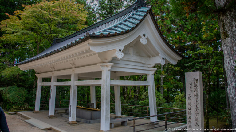 White open-sided purification pavilion over a stone basin beside an inscribed stone marker.