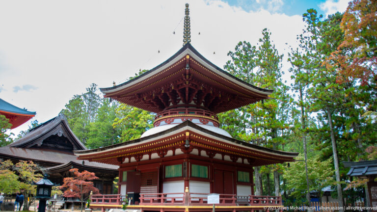 Corner view of a red-and-white two-story pagoda with layered roofs and tall finial among trees.