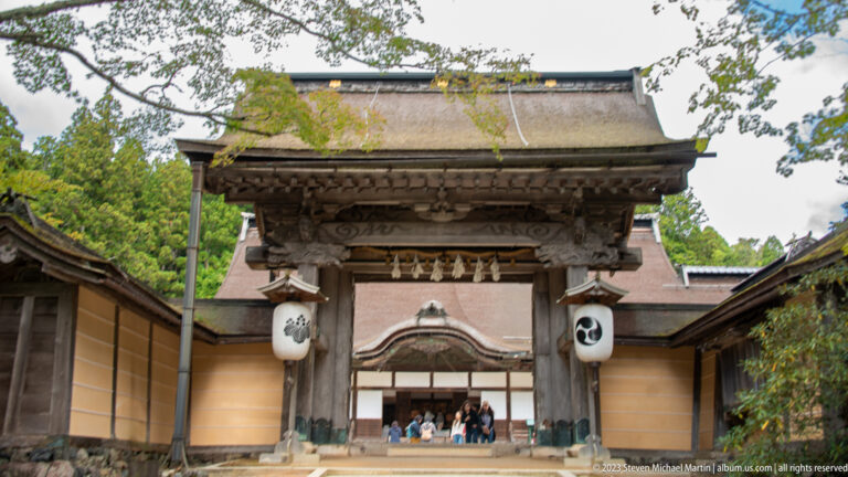 Weathered timber gate with cypress-bark roof, white crest lanterns, and ocher walls at Kongōbu-ji.