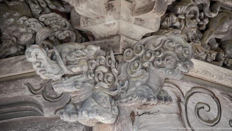 Close-up of a carved wooden shishi with open mouth and curled mane beneath a temple eave.