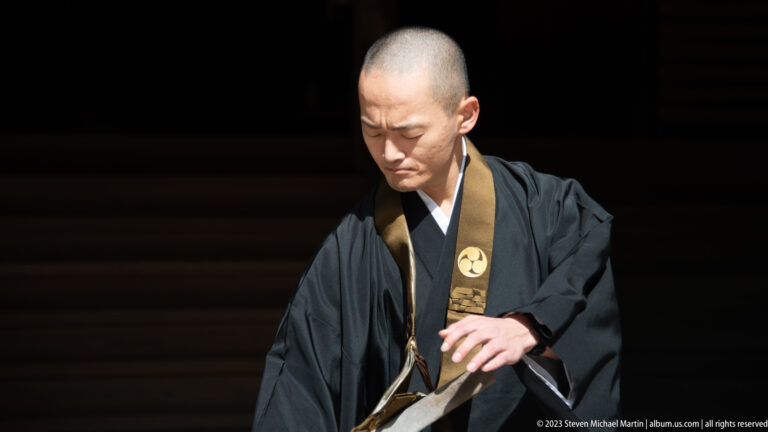 Shaved-head Buddhist monk in black robes and gold-brown stole looking downward against a dark background.