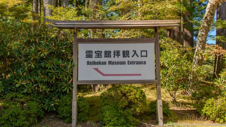 Wooden sign reading Reihokan Museum Entrance with a red arrow beside landscaped shrubs and trees.