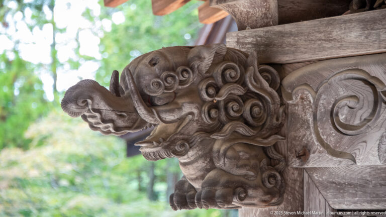 Close-up of a carved wooden shishi projecting from a bracket with green forest behind.