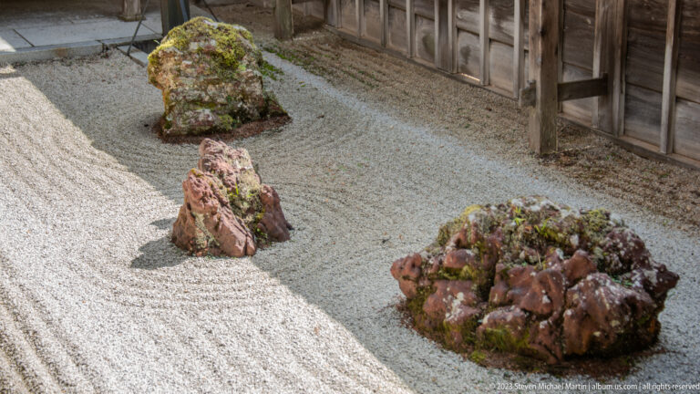 Reddish moss-covered stones arranged in raked white gravel beside a wooden temple corridor.