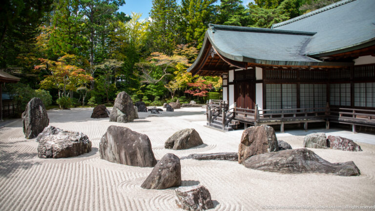 Jagged stones in raked white gravel beside a temple veranda and autumn trees.